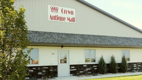 Exterior of the Crown Antique Mall, featuring a sign and a landscaped entrance with shrubs and a clear blue sky.