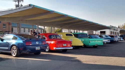 A row of classic cars in various colors parked under a shelter on a sunny day.