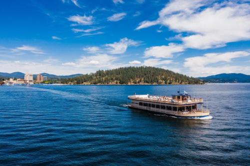 A boat cruises on a calm lake with a forested island and a city skyline in the background under a blue sky.
