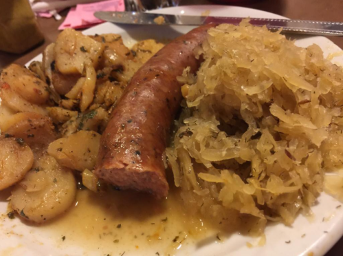 A plate featuring a sausage, sauerkraut, and seasoned potatoes, garnished with herbs and served with a knife and fork.