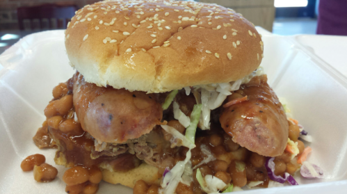 A sesame seed bun filled with sausages, barbecue sauce, baked beans, and coleslaw on a white takeout container.