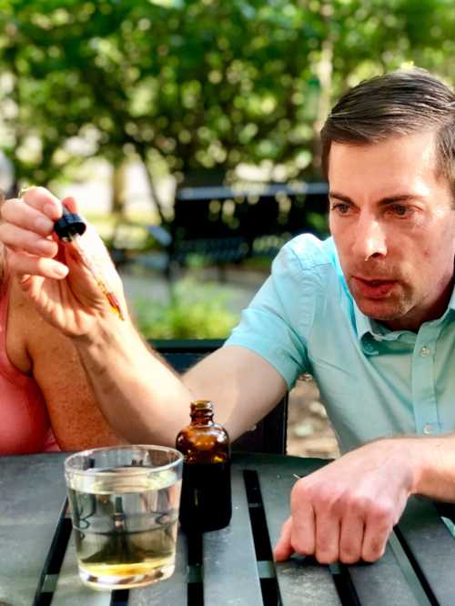 A man in a light blue shirt holds a dropper over a glass of water, focused on the task outdoors.