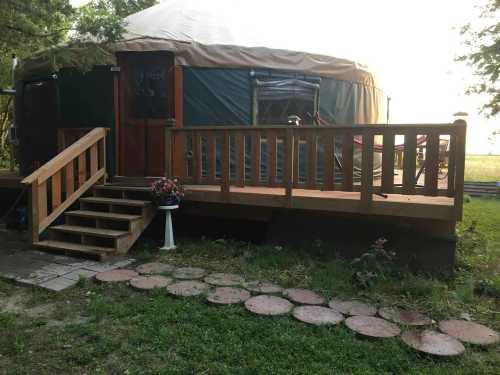 A wooden deck with steps leads to a yurt surrounded by grass and a stone path, with flowers in a pot nearby.