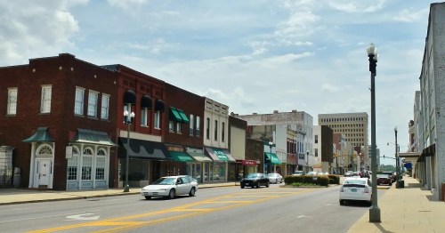 A street view of a small town with shops, cars, and a cloudy sky in the background.