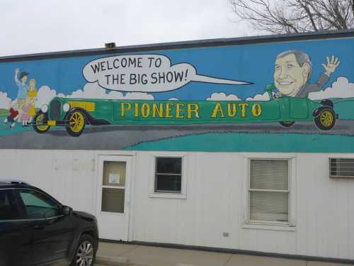 Mural on a building featuring a vintage car and a smiling man, with the text "Welcome to the Big Show!" above.