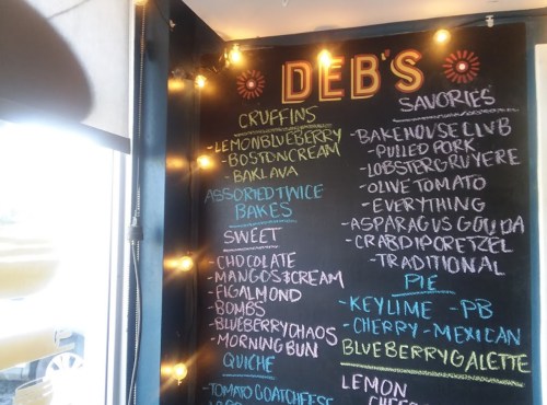 Chalkboard menu at Deb's featuring savory and sweet items like cruffins, bakes, and pies, decorated with string lights.