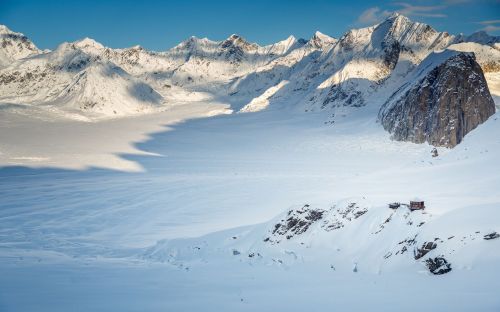 A vast snowy landscape with towering mountains and a small cabin nestled on a rocky outcrop.