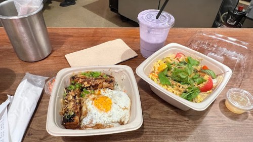 Two takeout containers on a wooden table: one with rice, grilled meat, and an egg; the other with noodles and vegetables.