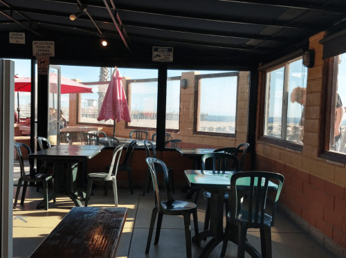 A bright, casual dining area with tables and chairs, featuring large windows overlooking a beach view.