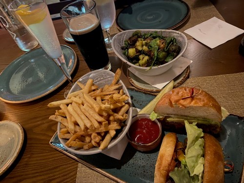 A table setting with two burgers, fries, Brussels sprouts, a cocktail, and a dark beer.