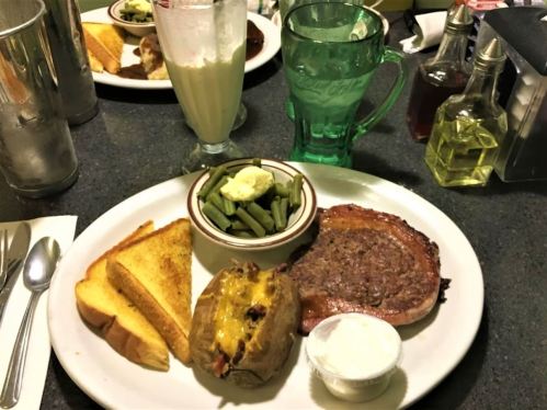 A plate with a grilled steak, baked potato with cheese, green beans, toast, and a milkshake on a diner table.