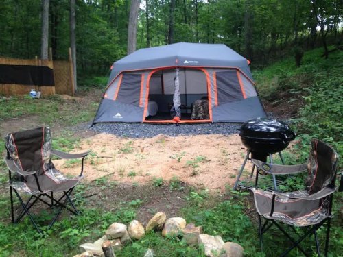 A camping scene featuring a large tent, two chairs, a grill, and a fire pit surrounded by trees and greenery.