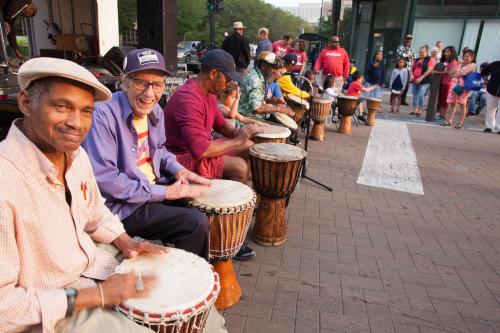 A group of musicians playing drums outdoors, with an audience watching in the background.