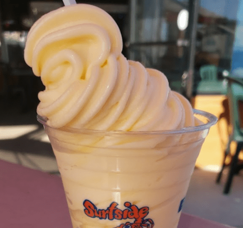 A tall swirl of creamy yellow soft serve ice cream in a clear cup, with a straw, against a bright background.