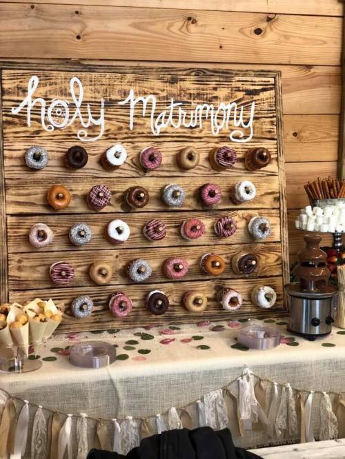 A wooden display wall with various donuts arranged, labeled "Holy Matrimony," alongside a chocolate fountain and treats.