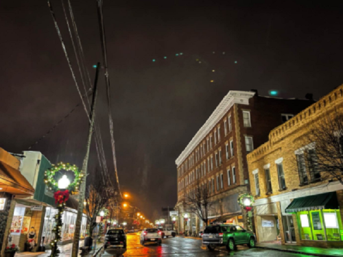 A quiet street at night, featuring festive lights, a historic building, and wet pavement reflecting streetlights.