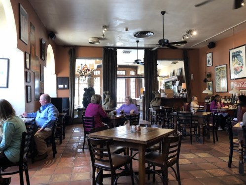 A cozy restaurant interior with wooden tables, patrons dining, and large windows letting in natural light.