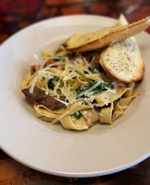 A plate of fettuccine pasta with vegetables, topped with cheese and served with two slices of garlic bread.
