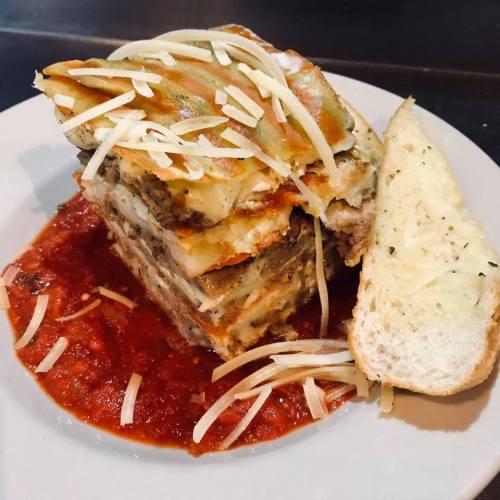 A layered lasagna topped with cheese, served on a plate with marinara sauce and a slice of garlic bread.