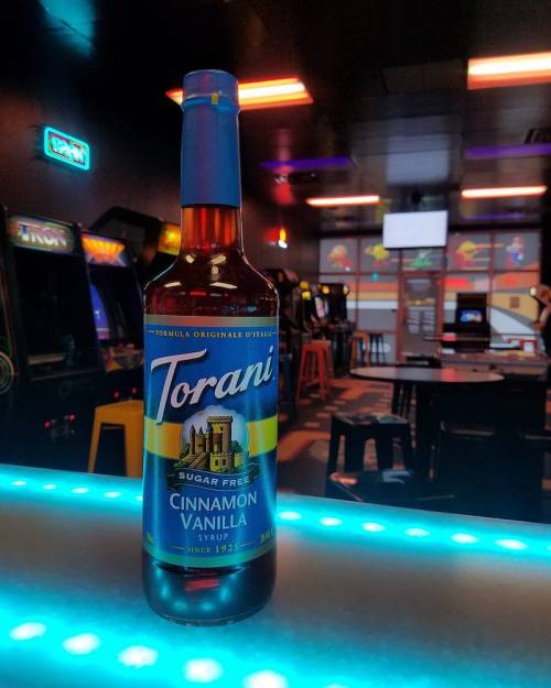 A bottle of Torani sugar-free cinnamon vanilla syrup on a bar counter, with arcade games in the background.