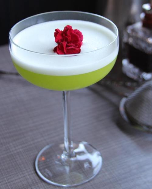 A vibrant green cocktail topped with a white frothy layer and a red flower garnish in a stylish glass.
