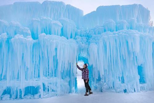A person in a plaid shirt stands near a large, icy blue ice formation with hanging icicles.