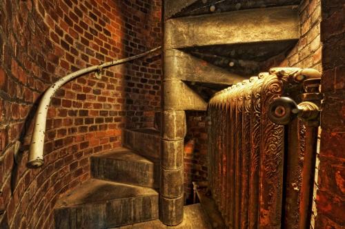 A spiral staircase made of stone and metal, surrounded by brick walls, with an ornate radiator at the base.