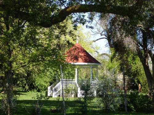 A white gazebo with a red roof surrounded by lush greenery and trees in a peaceful garden setting.