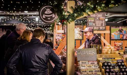 A festive market scene with people browsing stalls, lights twinkling overhead, and a vendor smiling at customers.