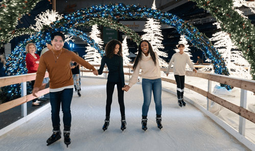 Four friends skate hand-in-hand on an ice rink decorated with festive lights and trees.