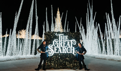 Two individuals pose beside a large illuminated sign reading "The Great Search," surrounded by festive lights and trees.