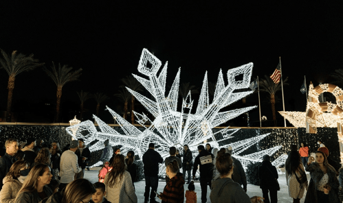 A crowd gathers around a large, illuminated snowflake display at night, with palm trees and festive lights in the background.