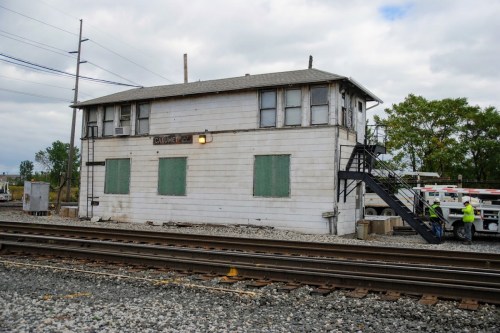 A weathered train control tower with green windows, surrounded by railroad tracks and workers in safety vests.