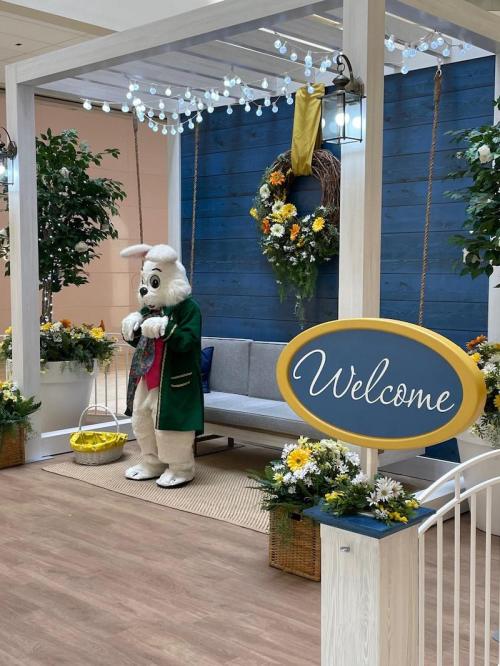A festive scene featuring a costumed bunny beside a "Welcome" sign, surrounded by flowers and decorative greenery.
