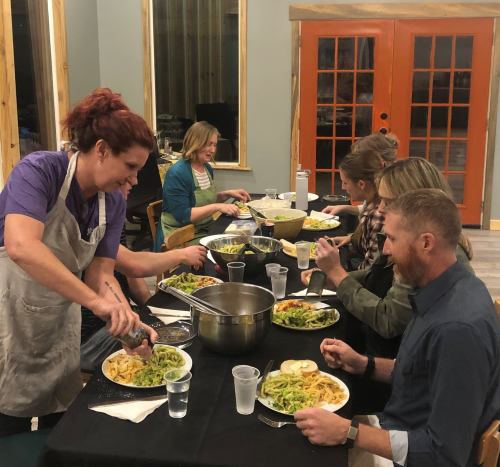 A group of people enjoying a meal together at a table, with one person serving pasta. Warm, inviting atmosphere.