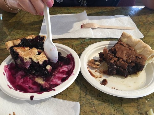 A hand holding a spoon next to two slices of pie: one blueberry and one chocolate, on paper plates.