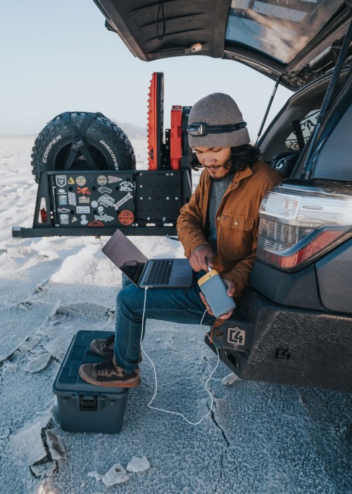 A person sits on a box by a car, using a laptop and charging a phone in a snowy landscape.