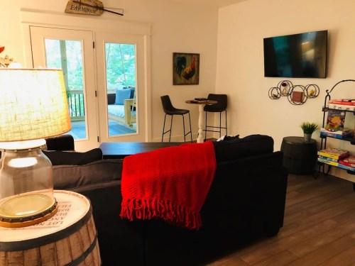 Cozy living space with a black sofa, red throw, lamp, and a TV, featuring a view of nature through glass doors.