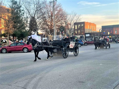 A horse-drawn carriage parade moves down a street lined with festive decorations and buildings.