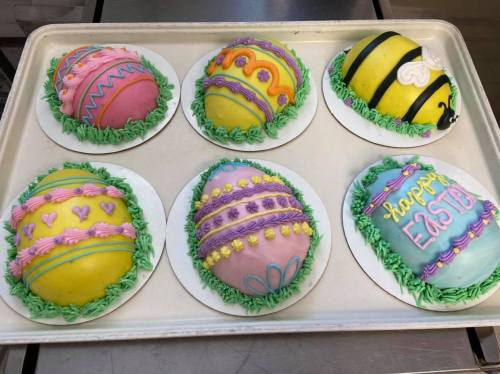 Six colorful Easter egg-shaped cakes decorated with icing and vibrant designs on a tray.