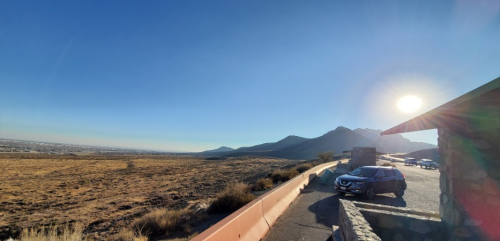 A panoramic view of a landscape with mountains, a clear sky, and a car parked near a stone structure.