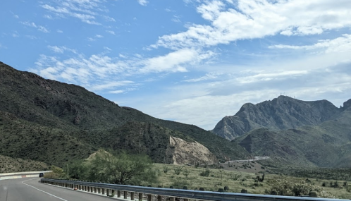 A scenic view of mountains and a clear blue sky along a highway, with greenery in the foreground.
