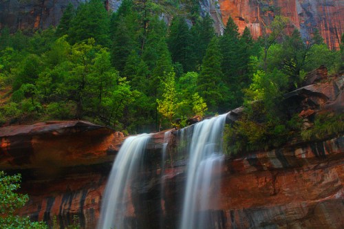 A serene waterfall cascades down rocky cliffs, surrounded by lush green trees and vibrant red rock formations.