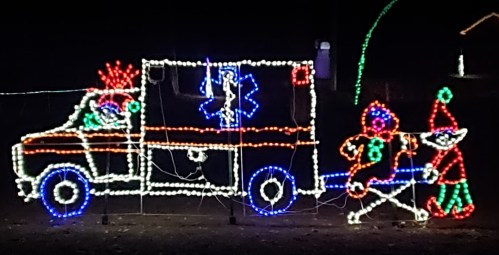 Colorful holiday lights depict an ambulance with festive characters, including a clown and an elf, on a stretcher.