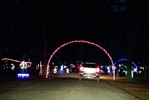A car drives down a dark road lined with colorful holiday lights and decorations in a festive display.