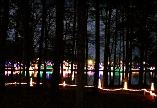 A serene night scene of a lake surrounded by trees, adorned with colorful holiday lights reflecting on the water.