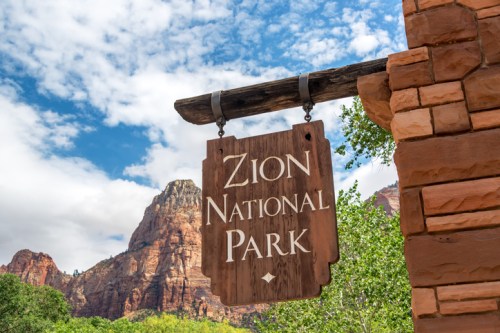 Wooden sign for Zion National Park against a backdrop of red rock formations and a blue sky with clouds.
