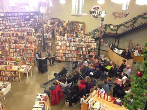 A cozy bookstore with shelves of books, featuring a reading event and an audience seated in front.