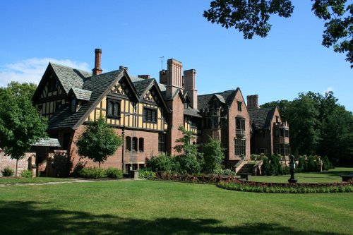 A large, historic mansion with a mix of brick and timber framing, surrounded by lush green lawns and trees.