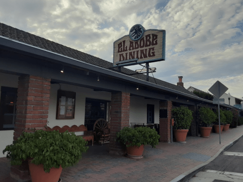 Exterior of Adobe Dining restaurant with a sign, brick columns, and large potted plants along the sidewalk.